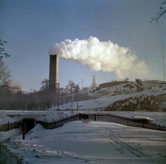 Chimney of a peak heating center carved into Alppila rock, seen from Vauhtitie, with Linnanmäki amusement park ferris wheel, Alppipuisto park, and modern water tower in the background