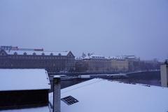 a scenic winter view of Charles Bridge in Prague with snow-covered rooftops and a partly cloudy sky