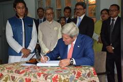 John Kerry speaking at a podium with Bangladeshi flags in the background