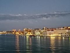 Tall Roman column seen from Brindisi port at night with lights and buildings.