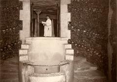 Guide with a hat and collection box at the entrance of the Paris Catacombs with large spiral staircase and walls covered with many skulls, 1929