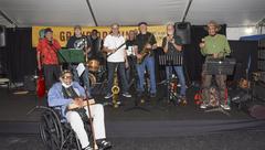 A nine men jazz band poses for a shot on stage. One man sits in wheelchair at the foot of the stage. The rest stand with various instruments including eclectic guitar, trombone, drums, saxophone, clarinet, trumpet, and a microphone.