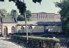 Sinebrychoffin puisto with a low courtyard building and a greenhouse in the foreground, and a brick building on the hill in the background