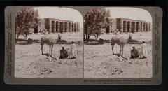 Camel and two people in foreground; man on donkey in front of ancient temple in Thebes, Egypt