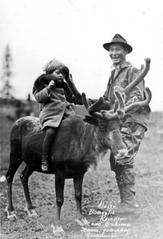 Eskimo man stands beside white child riding a reindeer in Anchorage, Alaska