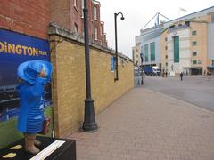 Paddington Bear statue in a sitting position wearing a blue coat and a hat, located at Stamford Gate