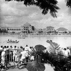 Crowds watching a rowing event by Takao River with damaged Kaohsiung Prefecture government building in the background