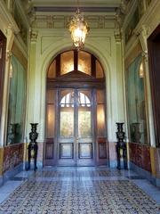 Vestibule of the Cousiño Palace with yellow, gray, and red marble panels, a distinctive bronze lantern at the top, Chinese vases from the Ming dynasty in the corners, Italian majolica floor, and Georges Clairin's oil paintings representing the four seasons with floral panels above them.