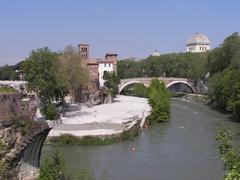 Southeast tip of Isola Tiberina viewed from Ponte Palatino with Ponte Quattro Capi connecting to the right and San Bartolomeo all'Isola tower in center-left. Ponte Rotto visible in far-left foreground.