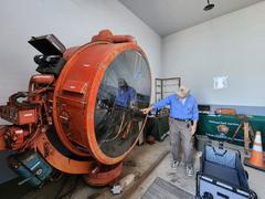 WWII military history volunteer pointing at a large 60 inch high-intensity carbon searchlight lens inside the gray-painted Searchlight Shed
