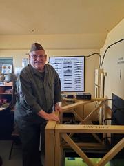 Military History volunteer in WWII-era green uniform leaning on yellow wooden railing guarding bunker ladder and beds