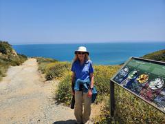 Park Rover volunteer standing by the Bayside Trail wildflower sign with yellow California sunflowers and blue San Diego Bay in the background