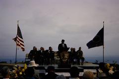 Man giving speech on stage at Cabrillo National Monument with NPS arrowhead on microphone stand