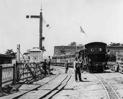 Standard gauge suburban train entering Cape Town station behind a 2-4-0T locomotive on dual gauge tracks with the Castle of Good Hope in the background