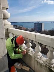 Person kneeling and scraping a sample from Belfry level exterior railing during inspection and lichen survey