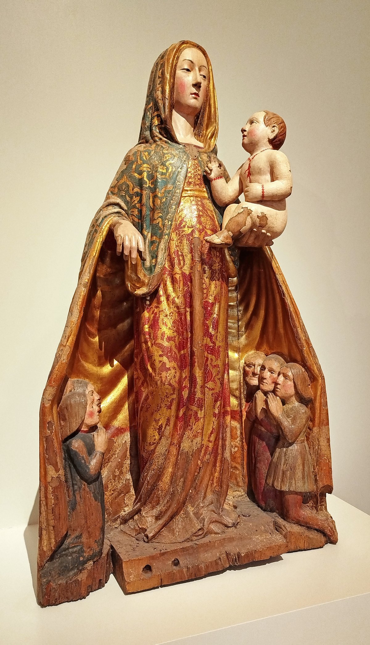 Wooden sculpture of Madonna della Misericordia holding baby Jesus and protecting praying faithful under her gold brocade cloak