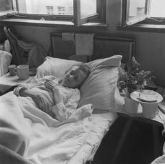 Woman lying on bed with drinking cups and a plate of bread beside her in Bergen-Belsen repatriation camp 1945