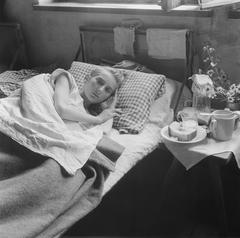 Woman lying on a bed with drink cups and a plate of bread beside her in Bergen-Belsen concentration camp after World War II repatriation
