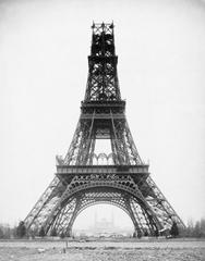 Black and white photograph of the Eiffel Tower under construction in 1888 showing its monumental iron lattice structure with a building dwarfed in the distance
