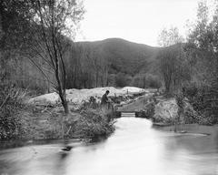 Black and white photograph of the Los Angeles River flowing through Griffith Park with a man in a bowler hat seated on the riverbank, trees and hills in the background, circa 1898-1910