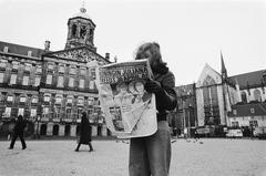 Young girl reading a morning newspaper with a bold headline about Queen Juliana's abdication on Dam Square, Amsterdam, with the Royal Palace on the left and the Nieuwe Kerk on the right, in black and white