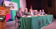 Six men seated at a table with emblems of Chamizal National Park and Mexico's Parque El Chamizal, with flags behind them, shaking hands during sister park signing ceremony
