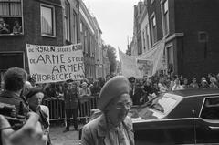 Queen Juliana arriving at Pieterskerk in Leiden with demonstrators behind holding a banner saying 'The rich getting richer, the poor getting poorer (Thorbecke 1844)'