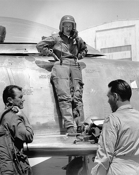 Jacqueline Cochran standing on the wing of her F-86 aircraft talking to Chuck Yeager and Bill Longhurst