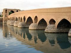 Old Shahrestan Bridge over water surrounded by trees under blue sky