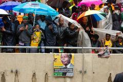 People in raincoats shield themselves with umbrellas during Nelson Mandela memorial service at FNB Stadium, South Africa