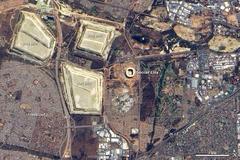 Aerial view of Soccer City Stadium in Johannesburg, South Africa, showing its renovated structure with high walls and long shadows, surrounded by Soweto Township and nearby slag heaps from gold mining.