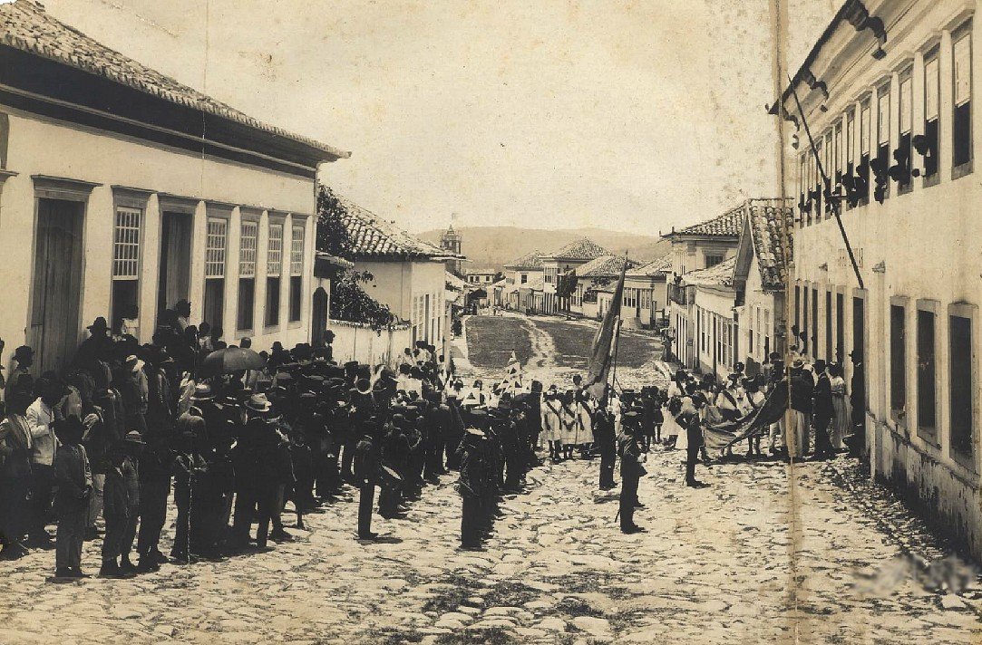 People observing an event with Brazilian and Minas Gerais flags in front of a school group and urban background