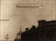 Airship flying above a city with tall buildings under a bright blue sky