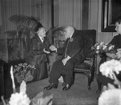 Actor Mari van Warmelo being congratulated by actress Rika Hopper at his 90th birthday reception in the foyer of Stadsschouwburg Amsterdam, 1956