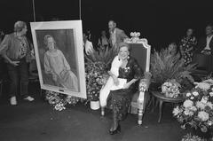Actress Ank van der Moer sitting on stage next to a large painted portrait of herself at Amsterdam Stadsschouwburg during her 50th theater anniversary celebration