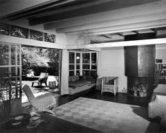 Interior view of the Rudolph M. Schindler House living room in West Hollywood, featuring period furniture, a fireplace, and sliding doors opening to a garden courtyard