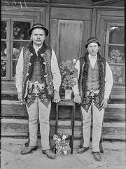 Two young Polish men in traditional Podhale costumes standing against a wooden cottage wall in Orava