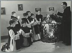 Monk conducting boys in religious robes singing Christmas carols by a nativity scene with candlelight in a Benedictine abbey school in Sint Andries, Belgium, 1935