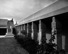 Exterior view of the Hollyhock House in Los Angeles featuring decorated pillars forming a colonnade, a row of plants, lawn, and marble staircase, designed by Frank Lloyd Wright in 1921