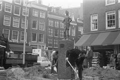 Historic black and white photo showing the Lieverdje statue being returned to Spui square in Amsterdam with the pit being filled, sculptor Carel Kneulman visible in the background