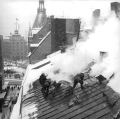 Firefighters cutting open the roof to extinguish attic fire at a warehouse building in central Stockholm, smoke billowing from the roof.