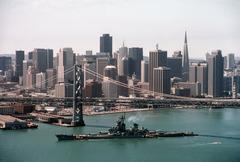 Aerial port beam view of the battleship USS New Jersey passing beneath the San Francisco Bay Bridge with San Francisco city in the background