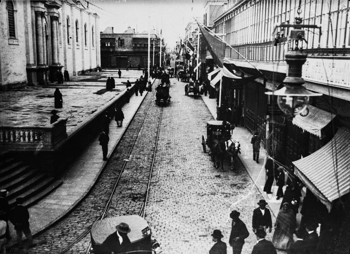 Street view of Calle de los Judíos (current Jr. Huallaga) next to the Lima Cathedral with hanging flags, pedestrians, horse-drawn carriages, commercial shops, and tram tracks