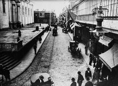 Street view of Calle de los Judíos (current Jr. Huallaga) next to the Lima Cathedral with hanging flags, pedestrians, horse-drawn carriages, commercial shops, and tram tracks