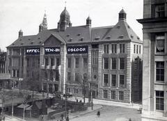 Historic black and white photo showing a building on Damrak in Amsterdam with white retouch paint spelling 'Effectenbioscoop' and a construction pit in front, indicating a fake April Fools' article about floor heating installation, Netherlands 1923
