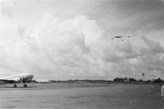 Black and white photo of a plane landing escorted by three Mustang aircraft over Kemayoran airport in Batavia, Indonesia on May 1, 1947