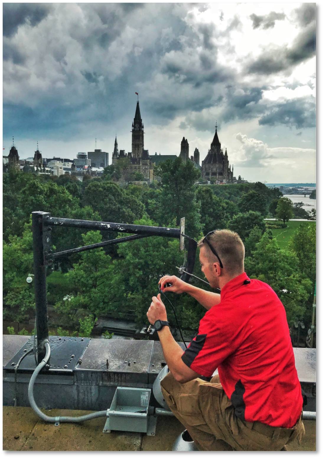 BU1 technician replacing a malfunctioning camera on the roof of the U.S. Embassy in Ottawa with Parliament Hill and Ottawa River in the background