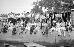 Crowd of people in street clothes gathered on the banks of Rivière-des-Prairies in Cartierville with boats and a megaphone