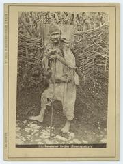 Bosnian Beggar in shabby clothes holding a stick barefoot in front of a wooden branch fence, early 20th century