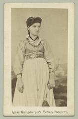 Studio portrait of a young unmarried Bosnian townswoman in traditional Christian urban clothing with a small vest, embroidered belt, cap, and jewelry against a mosque backdrop